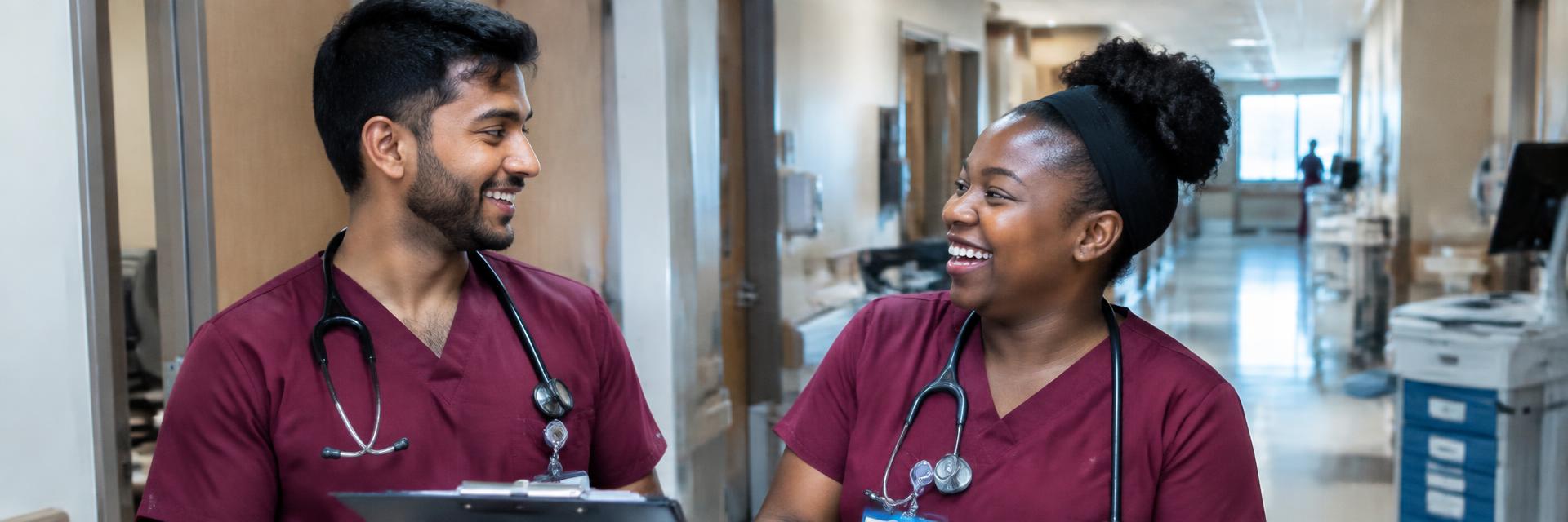 An Indian male nurse and a Black woman nurse in maroon scrubs laughing during a shift handoff